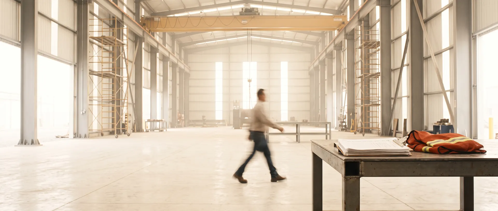 Nave de mantenimiento de una planta industrial bajo la luz de la mañana: grúa puente, andamios y una orden de trabajo sobre la mesa de acero